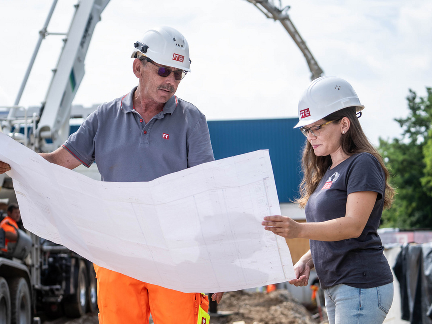 Zwei Mitarbeiter der Unternehmensgruppe Kassecker schauen gemeinsam auf einen Bauplan und stehen auf einer Baustelle.