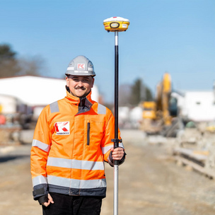 Ein Bauarbeiter in orangefarbener Sicherheitskleidung und Helm steht auf einer Baustelle und hält eine Vermessungsausrüstung. Im Hintergrund sind Baumaterialien und Baustellenfahrzeuge zu sehen.