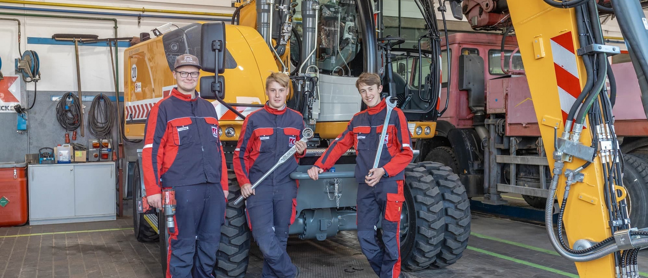 Drei Auszubildende in Arbeitskleidung stehen in einer Werkstatt vor einer großen Baumaschine und halten Werkzeuge in den Händen.