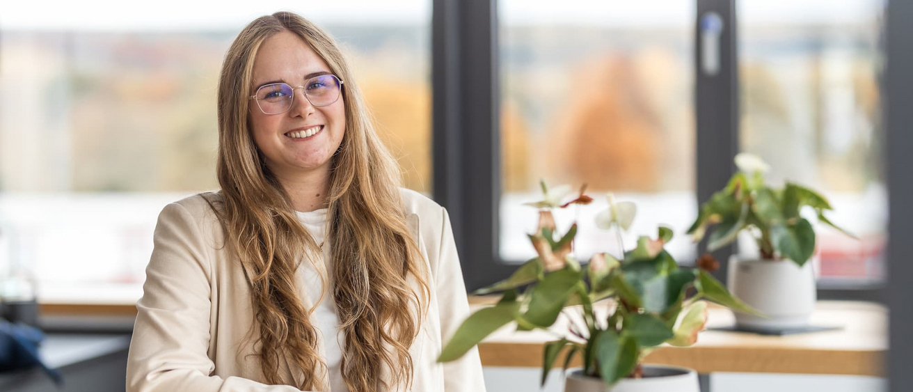 Eine lächelnde Frau mit langen blonden Haaren und Brille sitzt an einem Tisch im Büro der KASSECKER Gruppe, umgeben von Pflanzen. Sie trägt ein helles Jacket und ein weißes Oberteil.