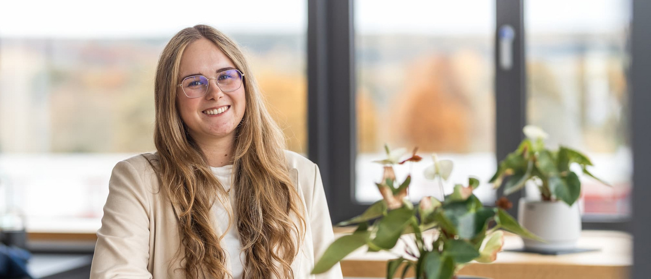 Eine lächelnde Frau mit langen blonden Haaren und Brille sitzt an einem Tisch im Büro der KASSECKER Gruppe, umgeben von Pflanzen. Sie trägt ein helles Jacket und ein weißes Oberteil.