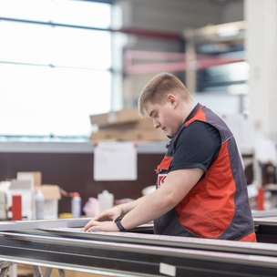 Ein junger Mann in Arbeitskleidung und Schutzweste arbeitet in einer Werkstatt an einem Metallrahmen. Im Hintergrund sind weitere Werkzeuge und Materialien zu sehen.