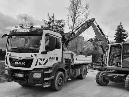 Ein LKW der Firma Sieber steht neben einem Bagger, der Erde in die Ladefläche des LKWs schaufelt. Im Hintergrund sind Bäume und ein Gebäude sichtbar.