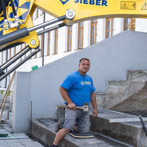 Sieber Mitarbeiter auf Baustelle Ein Mann kniet auf einer Treppe und hält einen Gummihammer. Im Hintergrund ein Bagger.
