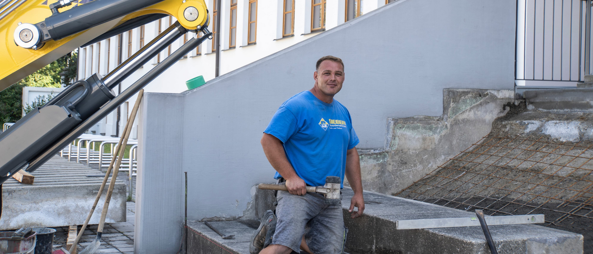 Sieber Mitarbeiter auf Baustelle Ein Mann kniet auf einer Treppe und hält einen Gummihammer. Im Hintergrund ein Bagger.