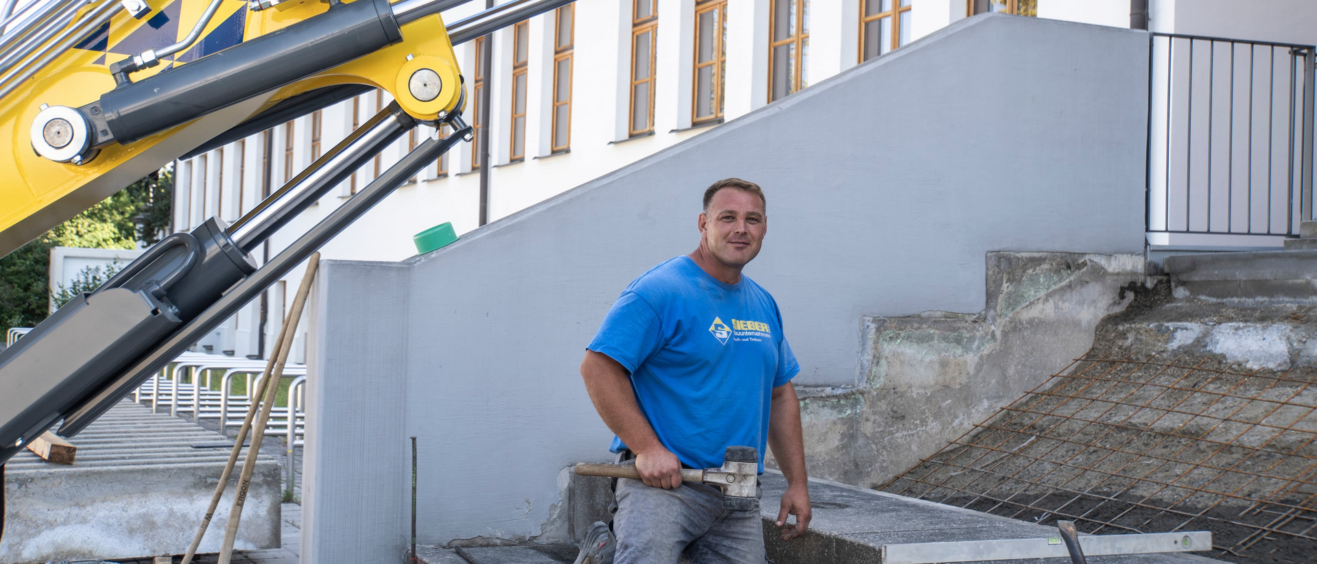 Sieber Mitarbeiter auf Baustelle Ein Mann kniet auf einer Treppe und hält einen Gummihammer. Im Hintergrund ein Bagger.