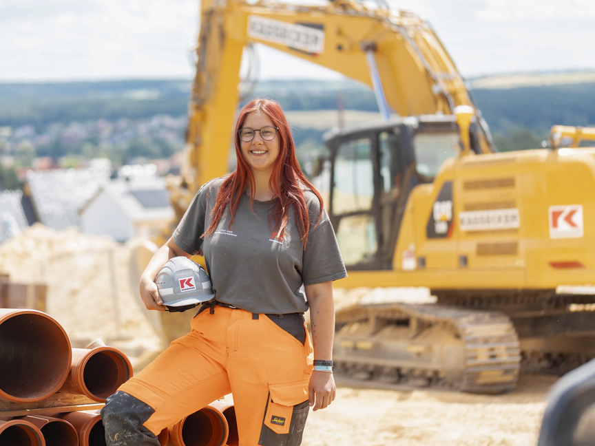 Eine junge Frau in Arbeitskleidung und Helm hält einen Bauhelm und steht neben großen Rohren auf einer Baustelle. Im Hintergrund sind Baugeräte und Erdhaufen zu sehen.