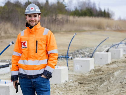 Ein lächelnder KASSECKER Mitarbeiter in orangefarbener Sicherheitskleidung und Helm steht auf einer Baustelle. Im Hintergrund sind Betonblöcke und Rohre zu sehen.