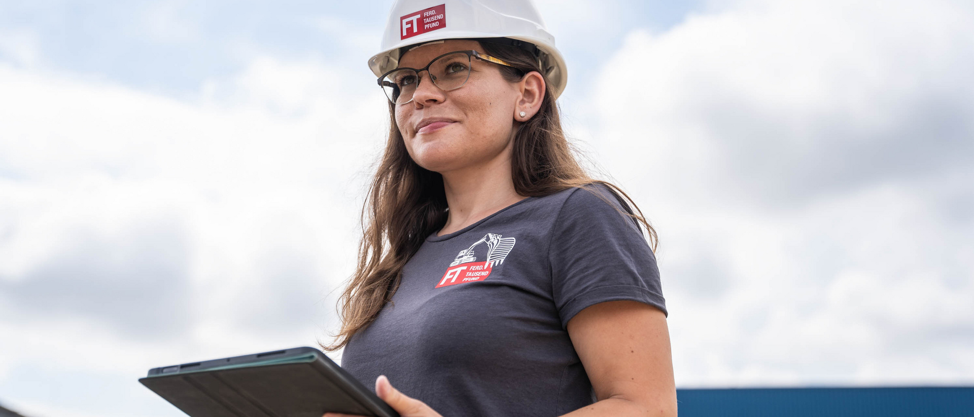 Bauleiterin der Unternehmensgruppe Kassecker Mitarbeiterin mit Bauhelm steht mit Tablet auf Baustelle und lächelt freundlich in die Ferne.