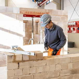 Ein Auszubildender in Arbeitskleidung und Helm mauert eine Wand in einer Werkstatt. Werkzeuge und Baumaterialien sind um ihn herum zu sehen.