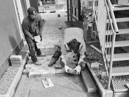 Zwei Bauarbeiter verlegen Pflastersteine auf einem Gehweg neben einem Gebäude. Eine Treppe ist rechts im Bild zu sehen.