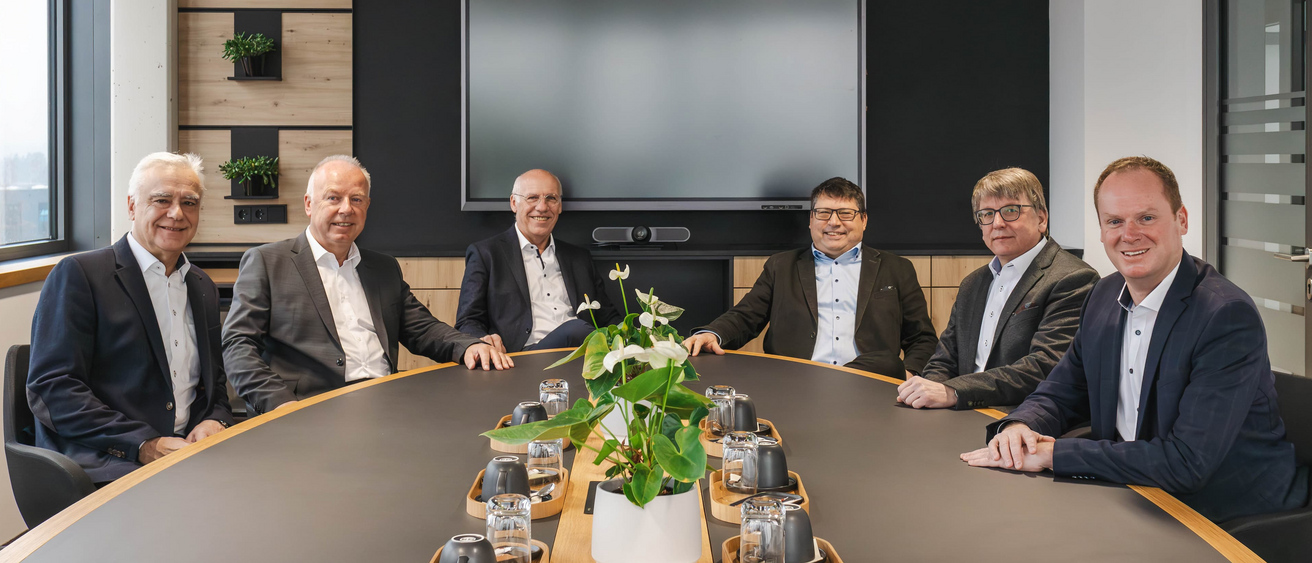 Sechs Männer in Businesskleidung sitzen an einem Konferenztisch in einem modernen Besprechungsraum. Im Hintergrund ist ein großer Bildschirm zu sehen.