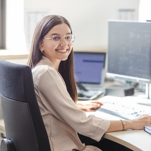 Eine junge Frau mit Brille sitzt an einem Schreibtisch in einem Büro und arbeitet an einem Computer. Sie lächelt in die Kamera.