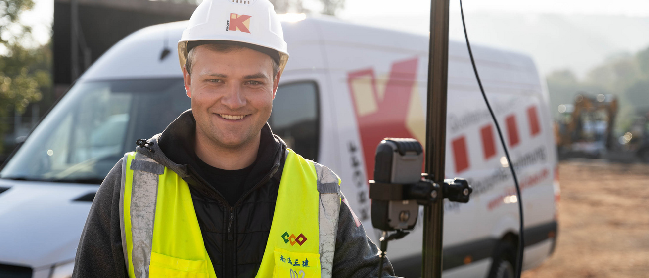 Junger Mann mit Vermessungsgerät blick freundlich in die Kamera. Er steht auf einer Baustelle, im Hintergrund ein Fahrzeug.