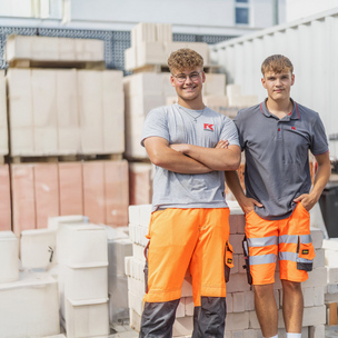 Zwei junge Männer lehnen an einem Haufen Ziegelsteinen und tragen Arbeitskleidung.