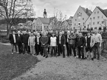 Eine Gruppe von Menschen steht draußen in einem Park und posiert für ein Gruppenfoto. Im Hintergrund sind Gebäude und eine Kirche zu sehen.