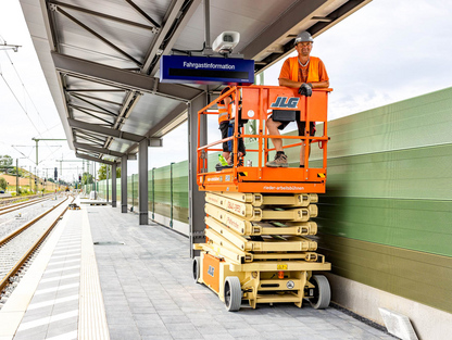 Ein Bauarbeiter auf einer orangenen Arbeitsbühne führt Arbeiten an einem Bahnsteig durch. Im Hintergrund sind Gleise und eine Überdachung zu sehen.