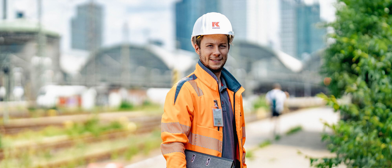 Ein Mitarbeiter der Unternehmensgruppe Kassecker steht mit einem Ordner in der Hand vor einer Baustelle in einer Stadt. Im Hintergrund sind moderne Hochhäuser und Bahnsteige zu sehen.