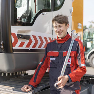 Ein junger Mann in Arbeitskleidung steht mit einem großen Schraubenschlüssel vor einer Baumaschine und lächelt in die Kamera.