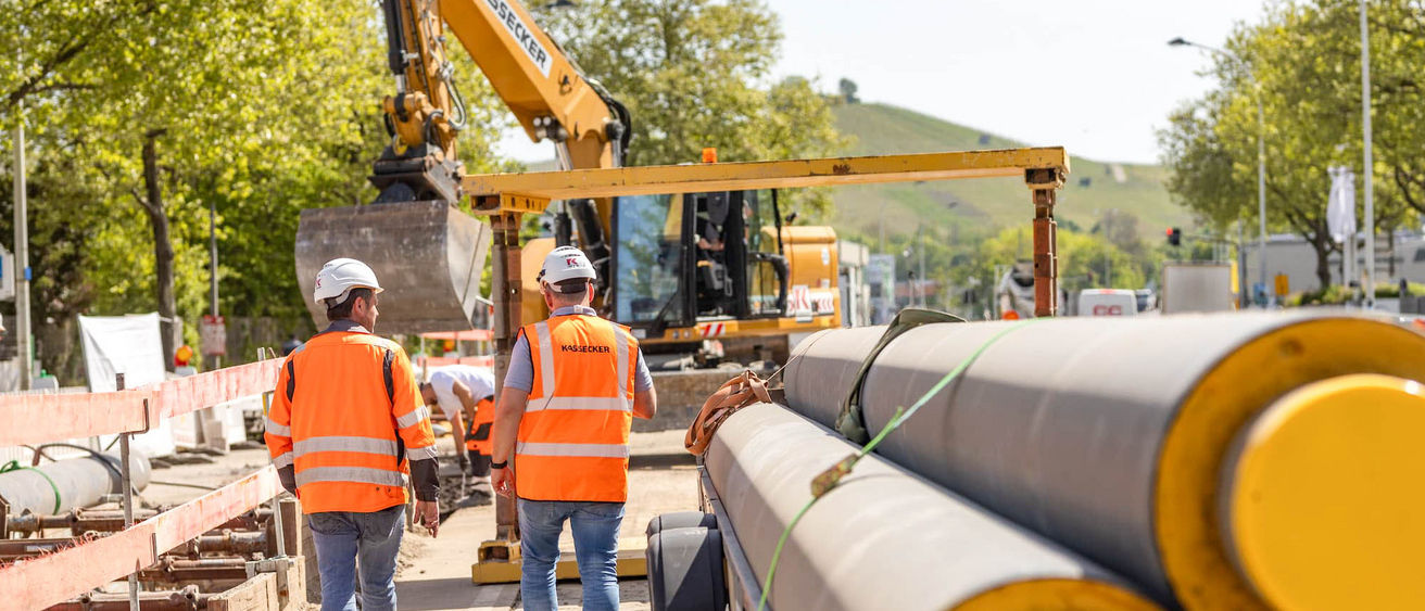 Zwei Bauarbeiter in Sicherheitskleidung und Helmen gehen auf einer Baustelle entlang einer Straße. Ein großer Bagger und Rohre sind im Hintergrund sichtbar.