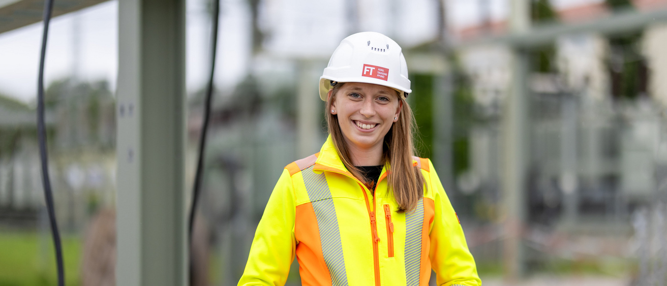 Freundlich lächelnde Mitarbeiterin von Tausendpfund steht auf Baustelle und trägt Arbeitskleidung und Helm.