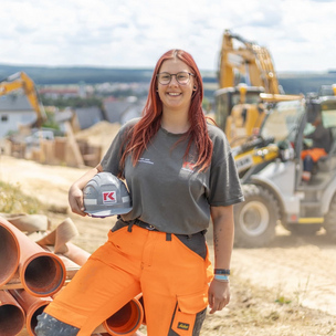 Eine junge Frau in Arbeitskleidung und Helm hält einen Bauhelm und steht neben großen Rohren auf einer Baustelle. Im Hintergrund sind Baugeräte und Erdhaufen zu sehen.