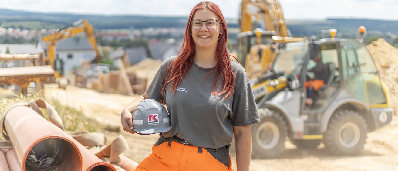 Eine junge Frau in Arbeitskleidung und Helm hält einen Bauhelm und steht neben großen Rohren auf einer Baustelle. Im Hintergrund sind Baugeräte und Erdhaufen zu sehen.