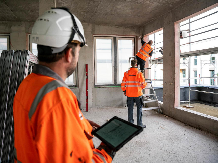 Bauarbeiter in orangefarbener Arbeitskleidung von KASSECKER arbeiten im Innenausbau eines Gebäudes. Einer der Arbeiter steht auf einem Gerüst, um Messungen vorzunehmen und ein anderer nutzt ein Tablet.