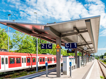 Ein moderner Bahnsteig mit Überdachung und Beschilderung. Im Hintergrund fährt ein roter Zug vorbei. Der Himmel ist klar mit wenigen Wolken.