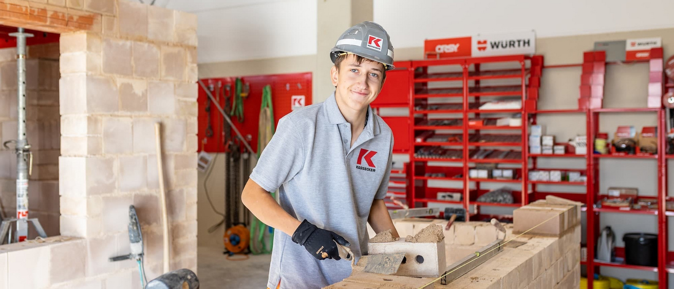 Ein Auszubildender Maurer in Arbeitskleidung und Helm arbeitet an einer Mauer in einer Werkstatt und lächelt in die Kamera. Im Hintergrund sind Werkzeuge und Baumaterialien zu sehen.