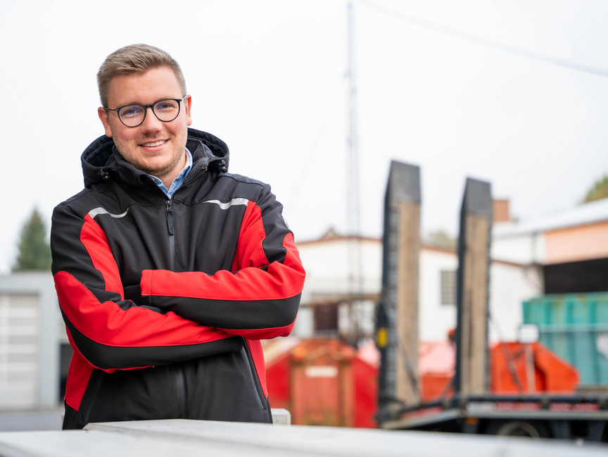 Freundlich lächelnder Mann mit Brille steht mit verschränkten Armen auf einem Bauhof.