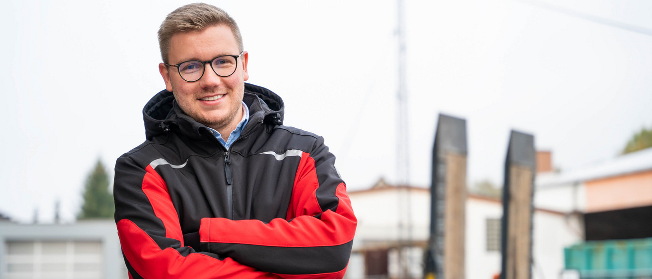 Freundlich lächelnder Mann mit Brille steht mit verschränkten Armen auf einem Bauhof.